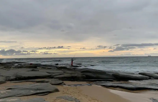 A lone fisherman stands on a rocky shoreline, casting their line into the ocean under an overcast sky.