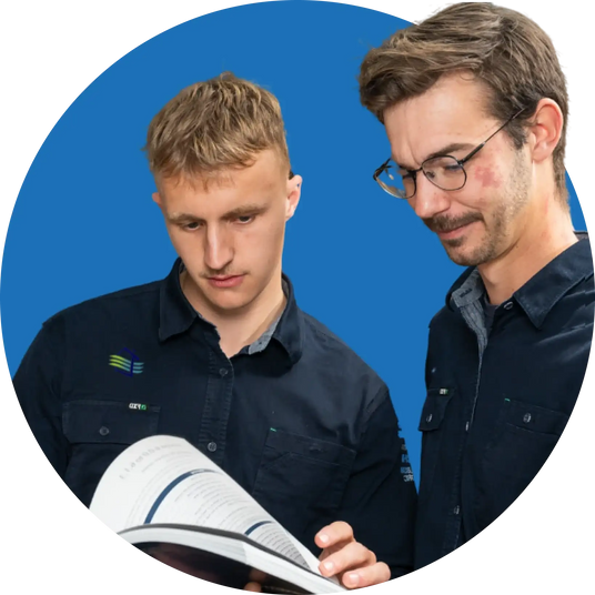 Two men in dark blue work shirts with subtle logos, one holding an open technical manual.