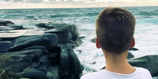 A young boy with short brown hair wearing a white t-shirt stands facing the ocean, his back to the camera.