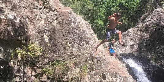 A young man in blue swim trunks leaps mid-air between rugged, mossy rocks near a cascading waterfall.